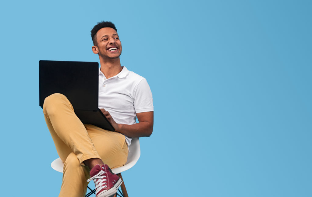 Young African American man smiling and looking at empty space while sitting on chair and using laptop against blue background